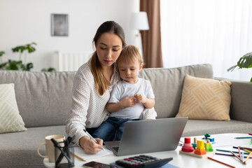 Motherhood and education. Young lady with baby on hands studying online on laptop and taking notes to workbook