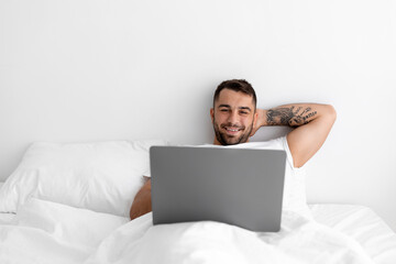 Satisfied relaxed millennial european muscular man lies on white bed and surfing in computer in bedroom