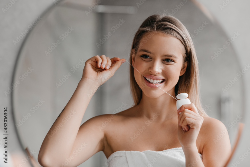 Wall mural Oral hygiene. Pretty lady brushing teeth with dental floss at home, standing in white towel and looking in mirror