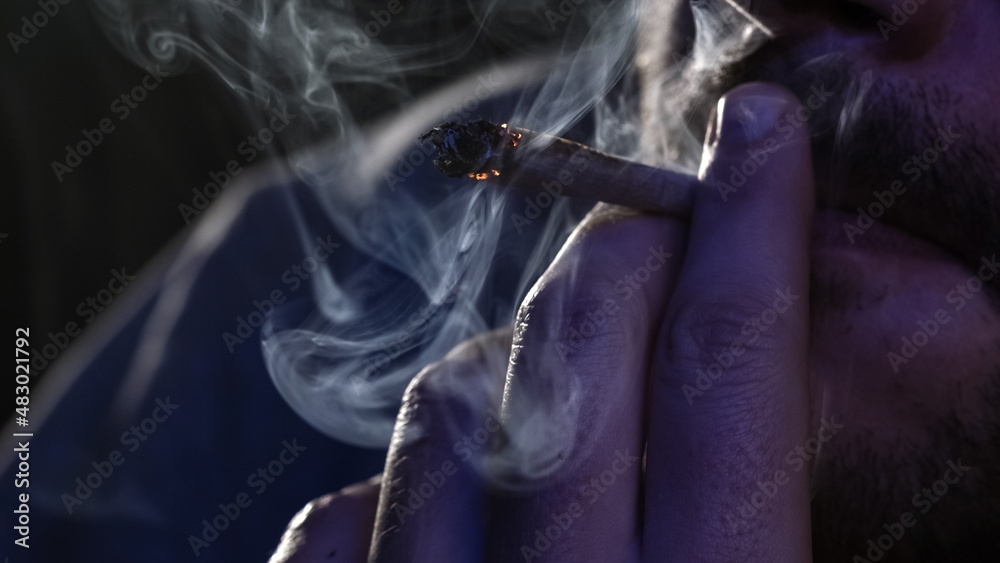 Wall mural Male fingers with burning cannabis cigarette with smoke and cigarette glow under the blinking light, macro view.