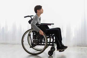 Full length portrait of impaired teenage boy sitting in wheelchair at home, side view