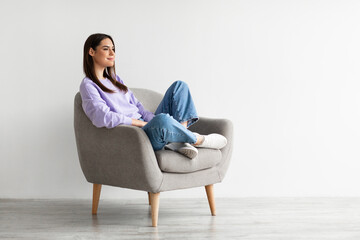 Young Caucasian woman relaxing in cozy armchair against white studio wall, copy space