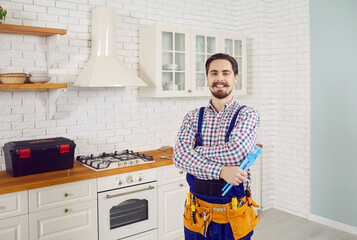 Portrait of smiling young plumber with tool in uniform pose at modern kitchen in client home. Happy...