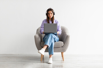 Young Caucasian woman having video call, using laptop computer, sitting in armchair, wearing wireless headphones - obrazy, fototapety, plakaty