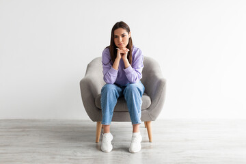 Full length of young woman sitting in armchair with dull or thoughtful face expression, having...