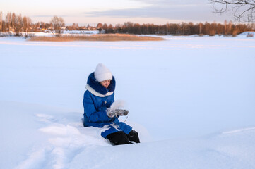 pretty girl in warm clothes is sitting in snow. Teenage girl walks on frosty morning and enjoys nature, holding snow in her palms