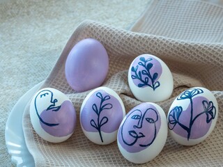 Beautiful Easter colored eggs on a kitchen towel, on a white plate, next to green sprigs of grass and leaves. The eggs are painted with a floral ornament and a human face in purple.