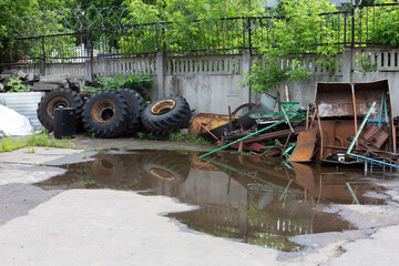 large old used truck wheels and tires in a pile next to metal sheets, bars and other rusty industrial rubbish in a dump corner with a puddle in front, a concrete wall and green trees in background