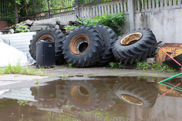 large old used rusty truck wheels and tires in a pile next to a concrete wall in a dump corner with a puddle in front