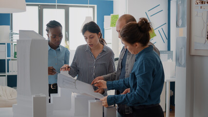 Diverse group of colleagues working on building model design with blueprints plan. Coworkers using maquette and industrial sketch to create architecture project, brainstorming ideas.