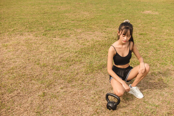 A slim and petite Filipino woman rests and kneels while looking at her kettlebell. At an grassy open field. Outdoor exercise lifestyle. - obrazy, fototapety, plakaty