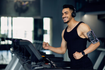 Fototapeta na wymiar Handsome Muscular Arab Man In Wireless Headphones Jogging On Treadmill At Gym