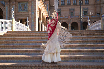 Obraz premium Young flamenco woman, Hispanic and brunette, in typical flamenco dance suit, dancing with red manila shawl with golden flowers, on stairs. Flamenco concept, dancer, typical Spanish dance.