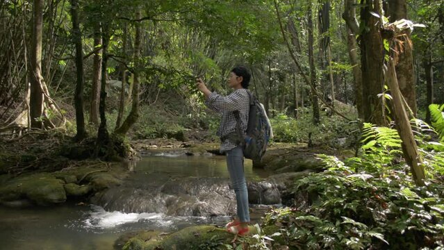 Asian female tourist with backpack standing on the rock near the water stream and enjoy taking picture in tropical forest during summer. Solo traveler. Outdoor pursuit. Jungle adventure in Thailand.
