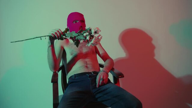 Anonymous shirtless man in balaclava sitting with flowers. Unrecognizable dangerous male with naked torso in pink balaclava sitting in chair near wall with bouquet of white flowers in hand
