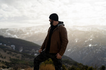 Chico joven con barba muy alto en la montaña con ropa marron y con montañas nevadas de fondo
