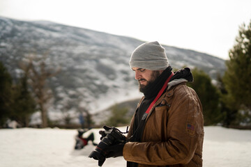 Chico con barba en la nieve con camara de fotos con ropa marron