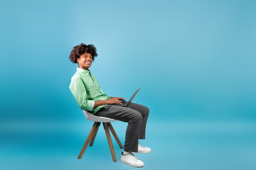 E-learning concept. Black guy student using laptop computer, sitting in chair over blue studio...
