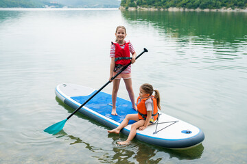 Girls padding on stand up paddle boarding on lake district. Children in swim life vest learning paddleboarding on SUP board. Family active leisure and local getaway with kids concept
