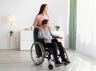 Full length of young mom and her impaired son in wheelchair looking out window together at home