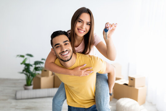 Cheerful Caucasian lady getting piggyback ride from her Arab boyfriend, showing house key indoors - Powered by Adobe