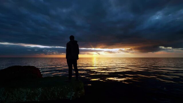 The silhouette of a man standing on a rock beside the water's edge watching the sun going down over the ocean horizon.