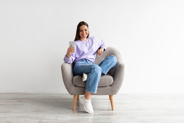 Smiling millennial woman chatting online on smartphone, sitting in comfy armchair on white studio...