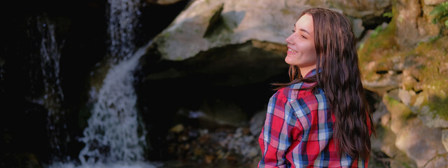 Young woman in white dress and plaid shirt enjoys Unity with nature outdoors on background of waterfall.