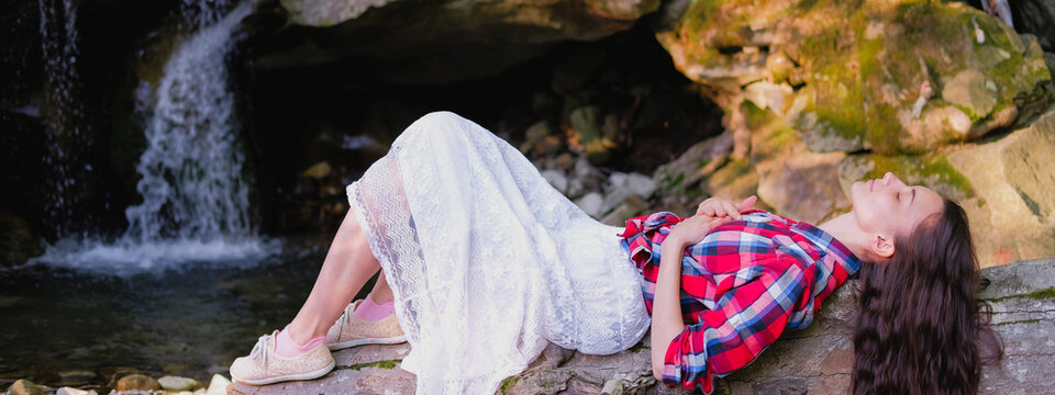 Young woman in white dress and plaid shirt lying on a log practicing breathing yoga pranayama outdoors on background of waterfall. Unity with nature concept.