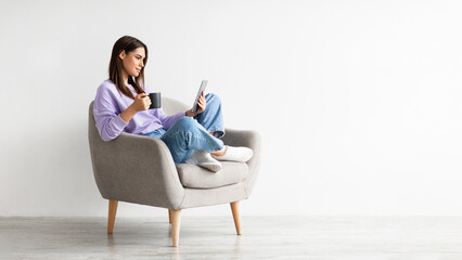 Young Caucasian woman with coffee sitting in armchair and using tablet computer against white studio wall