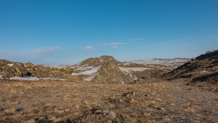 Snow-covered hills against a background of blue sky and clouds. Dry grass on stony ground. Winter Siberian landscape