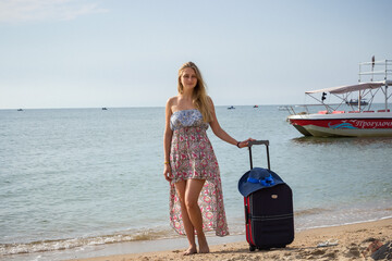 a young girl stands with a suitcase on a sandy seashore in a sundress and with a hat against the background of a boat