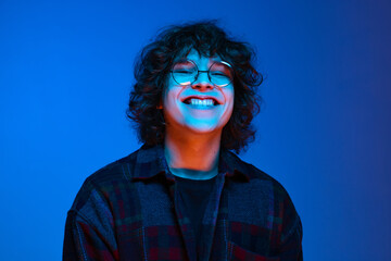 Portrait of young smiling man, student in black hoodie isolated on dark blue background in neon light. Concept of emotions, facial expression, youth, aspiration, sales - obrazy, fototapety, plakaty