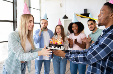Happy multiracial friends giving birthday cake with candles to excited young lady, having cool b-day party at home