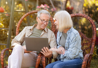 Fototapeta premium Happy elderly couple relaxing in wicker chairs in open air and using laptop computer, copy space