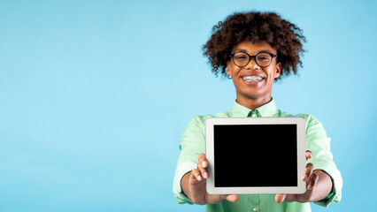 Cheerful black teen guy showing brand new digital tablet with blank screen over blue background, panorama, free space