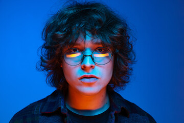 Close-up face of sad man, student looking at camera isolated on blue background in neon light. Concept of emotions, study, education, fashion