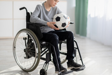 Disabled teen boy with soccer ball sitting in wheelchair, suffering over his injury, being unable to play game at home