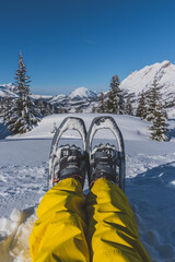 A first-person POV shot of a woman's legs in ski pants and snowshoes sitting during a hike in the French Alps on a cold winter day (L'Enclus, Devoluy, Hautes-Alpes)