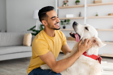 Happy young Arab man stroking his adorable dog in living room. Human animal friendship concept