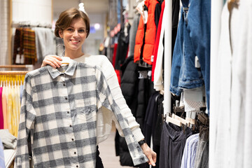 Fototapeta na wymiar Photo of young attractive brunette woman with a short haircut in a white sweater chooses stylish and casual shirt in a store in a shopping mall. Shop concept