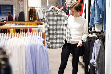 Photo of young attractive brunette woman with a short haircut in a white sweater chooses stylish and casual shirt in a store in a shopping mall. Shop concept