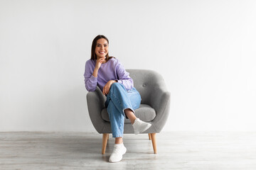 Full length of happy young woman in casual wear smiling at camera, sitting in comfy armchair...