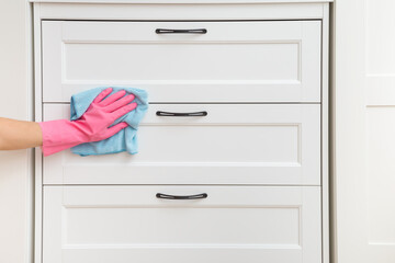 Young woman hand in pink rubber protective glove using blue dry rag and wiping dust from white wooden dresser surface in room. Closeup. Front view. Regular cleanup at home.