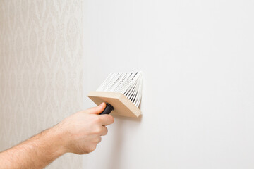Young adult man hand using brush and applying glue on white wall for wallpaper. Closeup. Repair...