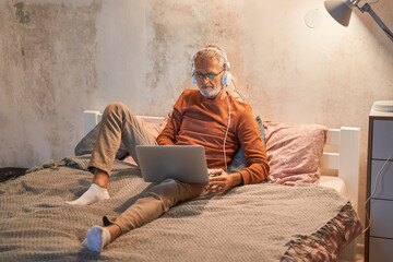 Senior man wearing headphones looking at the laptop while working with it on the bed