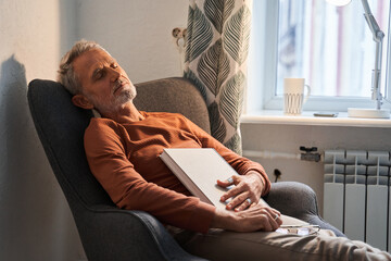 Tired mature man sleeping at the armchair with book on chest after reading - obrazy, fototapety, plakaty