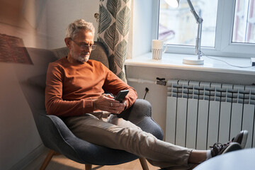 Carefree senior guy in domestic wear is sitting at cozy armchair