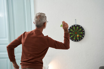 Mature caucasian man throwing darts in cozy room interior, while playing alone at home