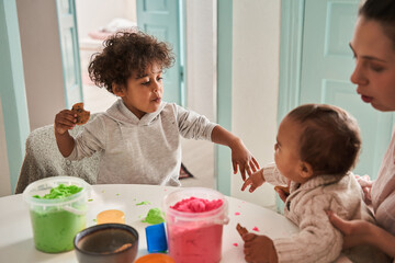 Two multiracial creative boys making figures from kinetic sand at the kitchen with their mother